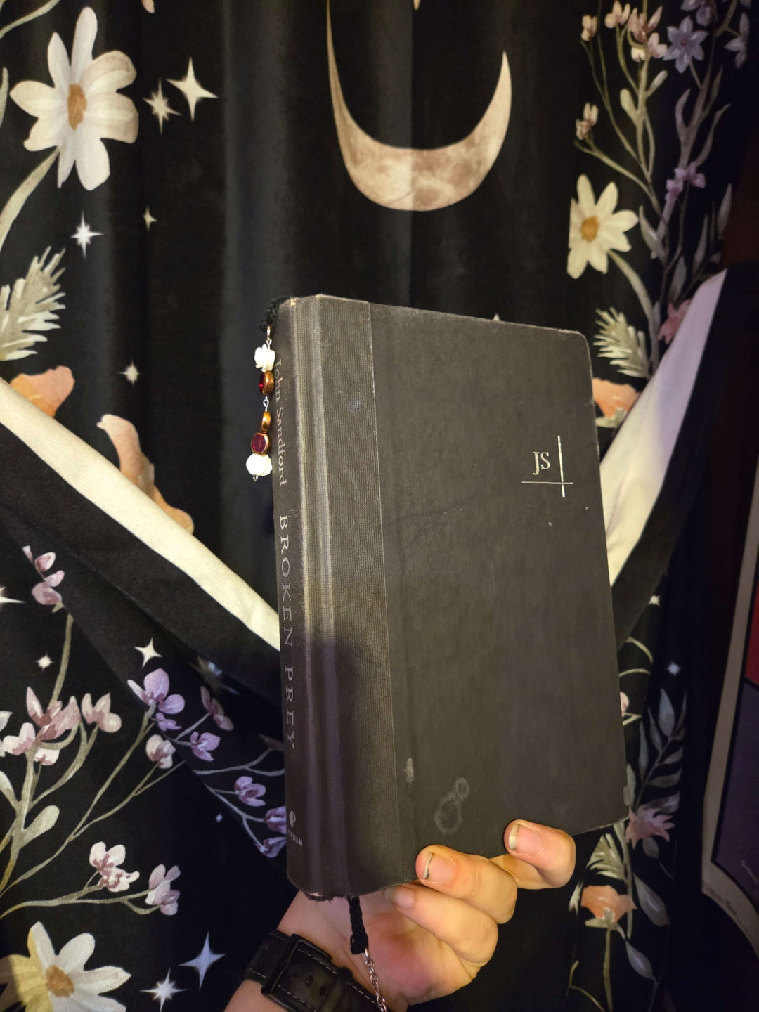 Bookmark displayed with a hand holding a book against a floral curtain background.